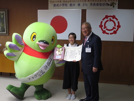 国旗と三郷市旗が吊り下げられている壁の前で、三郷市長と賞状と記念品の盾を手に持った鈴木奏さんがかいちゃんとともに記念撮影をしている写真