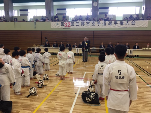 「第32回三郷市空手道選手権大会」と書かれた横断幕の下に並んだ長机の席に座った関係者と向かい合って立っている道着を着た選手たちが、中央に設置されたマイクスタンドの前で挨拶を行っている市長の話を聞いている様子の写真