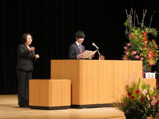 黒色のスーツを着た女性が手話を行っている右側の演壇でスーツ姿の新成人の男性が二十歳の主張を行っている様子の写真