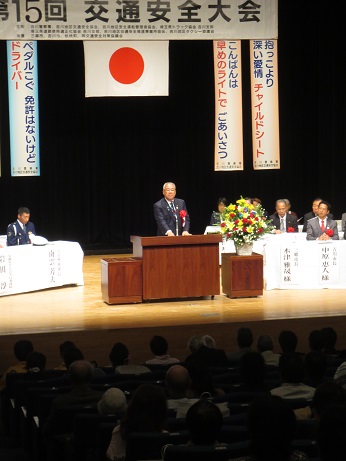「第15回交通安全大会」と書かれた横断幕の下に、左から「ペダルこぐ 免許はないけど ドライバー」の標語、国旗、「こんばんは 早めのライトで ごあいさつ」と「抱っこより 深い愛情 チャイルドシート」の標語が並んで掛けられたステージの、赤・黄色・白の豪華な花々で彩られた花台と並ぶ演台に立ち挨拶の言葉を述べる三郷市長と、ステージ上に配置された席に座って話を聞く来賓者の写真