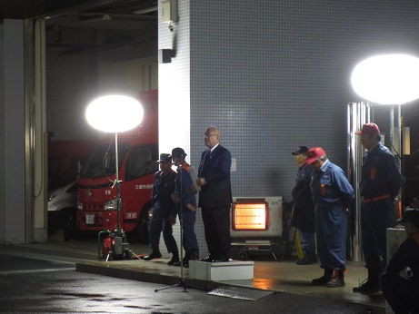 バルーンライトが設置された夜の消防署前で、踏み台の上に立ち挨拶をする市長と、その後ろに立つ数名の制服を着た消防団員たちの写真