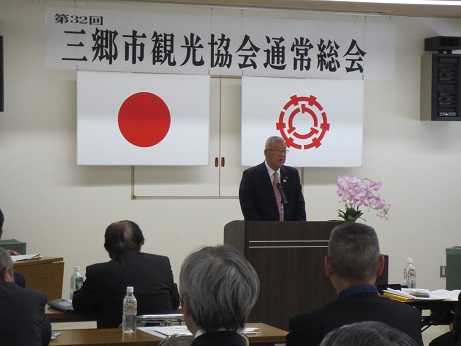 「三郷市観光協会通常総会」と書かれた横断幕と国旗・三郷市旗が吊るされている壁の前で演台に立ち話をする三郷市長と、ペットボトルの水と資料が置かれた席に座り話を聞く参加者の写真