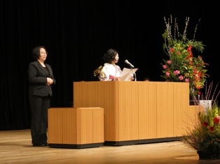 黒色のスーツを着た女性が両手を前に組んで立っている右側の演壇で振り袖姿の新成人の女性が二十歳の主張を行っている様子の写真