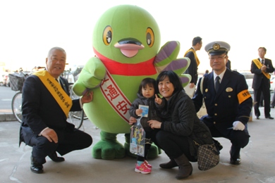 キャンペーングッズを貰った女の子と母親がマスコットキャラクターかいちゃんの傍で市長と警察署長の間に座り記念撮影している写真