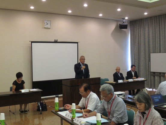 プロジェクタースクリーンの用意された部屋で、長机の資料を見ながら着席している関係者たちと、正面の演壇に立って話をしている市長の写真