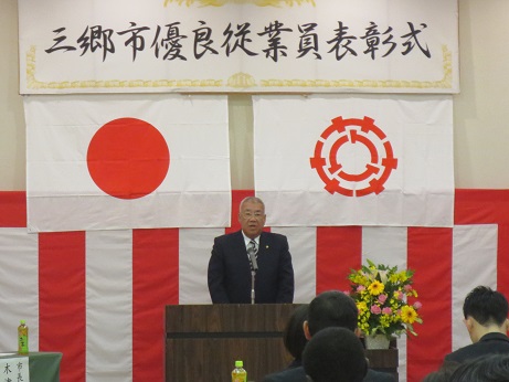 「三郷市優良従業員表彰式」と書かれた横断幕と国旗と三郷市旗、紅白幕が貼られた会場の檀上で挨拶を行っている市長の写真