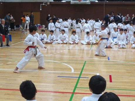 赤の帯を締めた子どもと黄色の帯を締めた子どもが形試合を行っており、あぐら座りをしている子どもたちがその様子を観ている写真