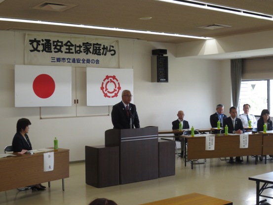 部屋の壁に「交通安全は家庭から」と書かれた紙が貼ってあり、両脇で関係者たちが座っている中、演壇に立って話をしている市長の写真