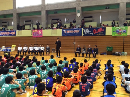 左右に設置された椅子に関係者が座り、中央に立つ山本選手の話を体育座りをしている子供たちが聞いている後ろ姿の写真