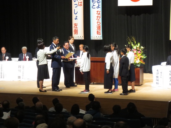 ステージ上の演台の前で表彰状の内容を読み上げる警察関係者とマイクを持ちサポートする女性、向かい合って立ち話を聞く第52回交通安全子供自転車埼玉県大会で8位入賞を果たした子どもたちの写真