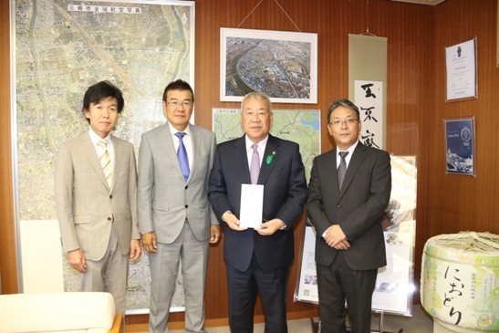 大きな航空写真などが飾られた室内で、頂いた義援金を持っている市長と3名の男性が横一列に並んでいる写真