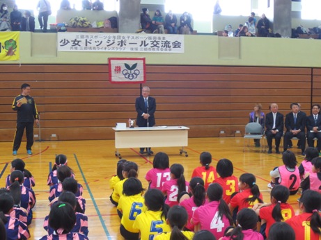 「少女ドッジボール交流会」と書かれた横幕が貼られた体育館で挨拶を行っている市長の話を、向かい合って座って聞いている子供たちの後ろ姿の写真