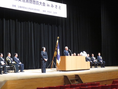 舞台の両脇に来賓の男性たちが座り、その中央手前に設けられた演台に立って客席を向いて話をする市長と、市長と少し間隔を空けて立っているスーツ姿の女性を写した挨拶の様子の写真