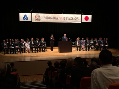 「三郷町制施行50周年記念式典」と書かれた横断幕の下のステージの左右に設置された椅子に座っている関係者や観客席に座っている参加者が、演壇に立ち挨拶を行っている市長の話を聞いている写真