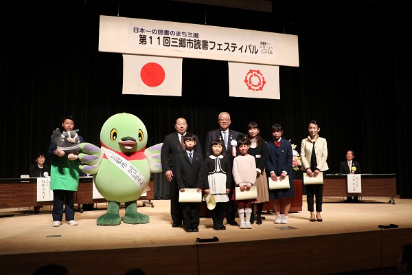 「第11回三郷市読書フェスティバル」と書かれたボードと国旗・三郷市旗が吊るしてあるステージ上で、表彰状を持った市長賞受賞者である子どもたちと女性が三郷市長と白いタスキをかけたかいちゃん、男子学生に抱えられたつぶちゃんとともに記念撮影をしている写真
