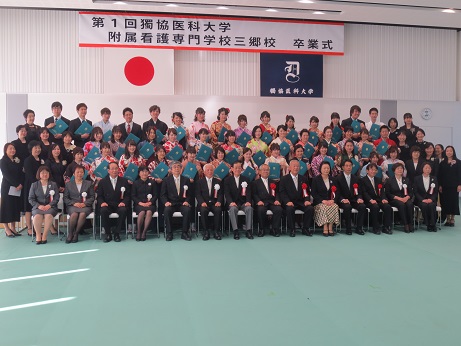 「第1回獨協医科大学附属看護専門学校三郷校卒業式」と書かれた横断幕と国旗・獨協医科大学の校章機が吊り下げられている壁の前で、三郷市長と卒業証書を持った袴姿の卒業生、関係者が大勢並び記念撮影をしている写真