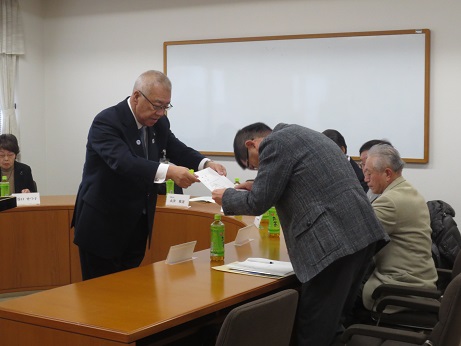 U字の会議用テーブル越しに、委嘱書を手渡す三郷市長と席札とペットボトルのお茶が置かれている席を立ちお辞儀をしながら受け取る男性の写真