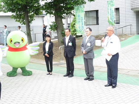 「エコ実践中緑のカーテン」と書かれたのぼり旗が2本設置されている市役所前のスロープのそばで みどりのカーテン定植式の挨拶の言葉を述べる三郷市長と、横に並んで話を聞く職員3名とかいちゃんの写真