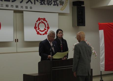 演台に立って表彰状を読み上げている市長と、その前で姿勢を正して立っている男性、市長の後方に控える女性職員の写真