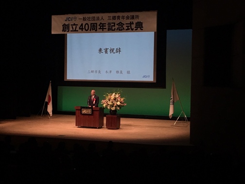 「一般社団法人三郷青年会議所創立40周年記念式典」と書かれた横断幕の下に設置されたスクリーンに「来賓祝辞」と表示され、ステージの中央に設置された演壇で挨拶を行っている市長の写真