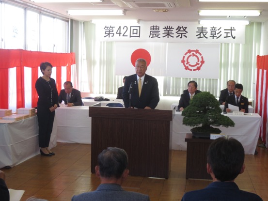 「第42回農業祭表彰式」と書かれた横断幕と国旗・三郷市旗が吊り下げられている会場で、立派な盆栽が置かれた花台と並ぶ演台に立ち挨拶の言葉を述べる三郷市長と、後ろに配置された白い布が掛けられ席札が置かれている席に座り話を聞く関係者の写真