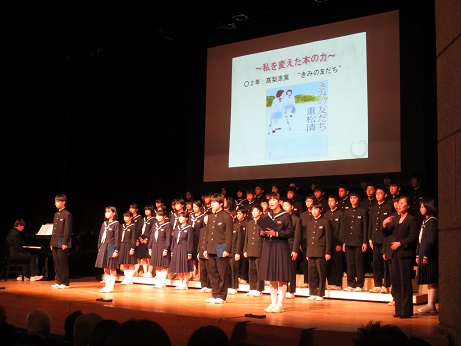 舞台上で、取り組み発表のスライドが映し出された後方のスクリーンの前に、制服を着たたくさんの中学生が前後5列に並び正面を向いて立っている発表の様子写真