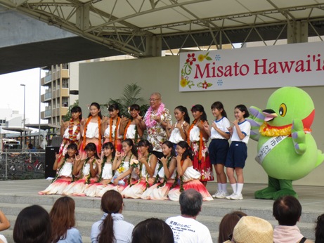 マスコットキャラクターかいちゃんとアロハシャツを着た市長を中央に白色とオレンジ色の衣装を身に着けた女子生徒達や体操着を着た女子生徒が2列に並んでいる記念写真