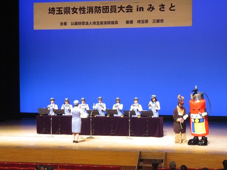 「埼玉県女性消防団員大会inみさと」と書かれた看板が掲げられた舞台の上でハンドベルを演奏する制服を着た女性消防団員たちと、その横に立つ消化器のマスコットの写真