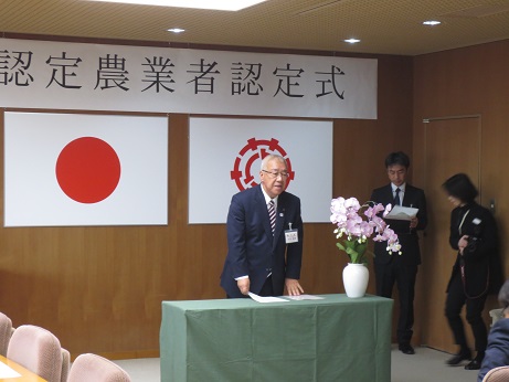 「認定農業者認定式」と書かれた横長のパネルと、国旗、三郷市の旗が張られた壁を背に、演台に立って挨拶をする市長の写真