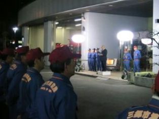 左右に大きな照明が照らされているなか、小さな台の上に立っている市長の話を消防団員の方々が整列して並び聞いている写真