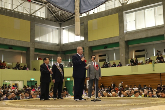 土俵に3名の男性と、中央に設置されたマイクスタンドの前で挨拶を行っている市長の写真