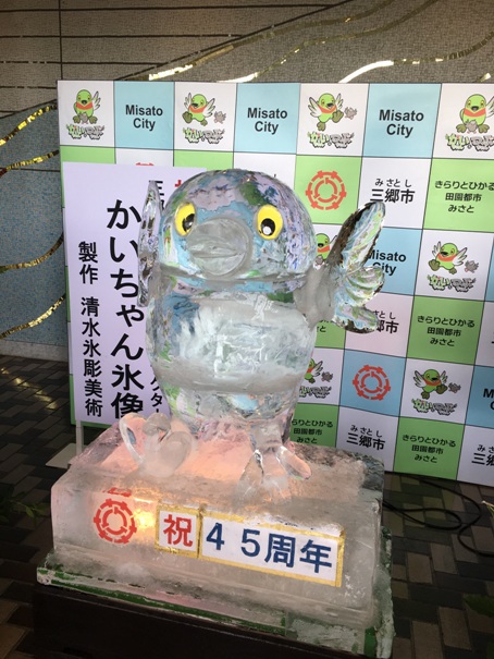 三郷市のバックボードの前に設置され、三郷市のロゴと「祝45周年」の文字が書かれた氷の土台の上にかいちゃんが手を広げて立っている清水氷彫美術が製作したかいちゃん氷像の写真