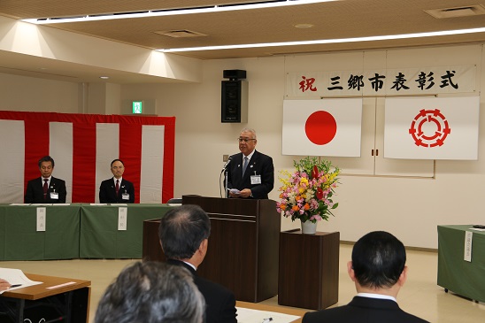「祝三郷市表彰式」と書かれた横断幕が貼り付けられ国旗・三郷市旗が吊るしてある壁の前で、赤・ピンク・黄色・オレンジ・白の豪華な花々で彩られた花台と並ぶ演台に立ち挨拶の言葉を述べる三郷市長と、席に座り話を聞く受賞者の写真