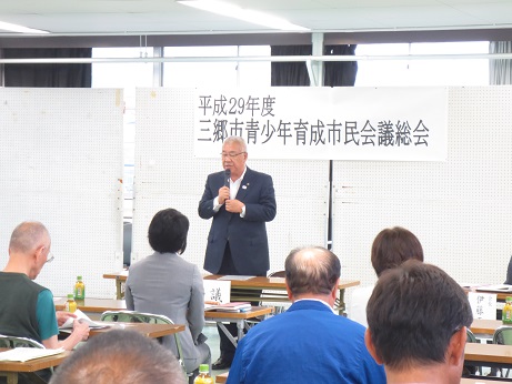 「平成29年度三郷市青少年育成市民会議総会」と書かれたポスターが貼りつけられた白いボードの前で、あいさつの言葉を述べる三郷市長とペットボトルのお茶が置かれた席に座り話を聞く参加者の写真