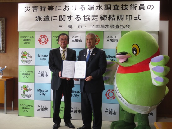 三郷市のバックボードに「災害時等における漏水調査技術員の派遣に関する協定締結調印式」のボードが設置されている部屋で、市長と古賀会長が協定書を一緒に持っている横にかいちゃんが立っている写真