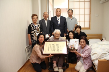 車いすに座って額縁に入った賞状を持った高橋たけ子さんの周りに7名の関係者と市長が並んでいる記念写真
