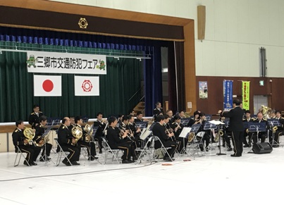 舞台を背に扇形に置かれた椅子に座って管楽器を演奏する制服姿の埼玉県警察音楽隊と指揮者を左斜めから写した写真