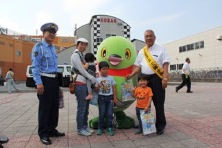 交番前の広場で警察署長と市長が立っている間に、2名の男の子と小さな子供を抱っこした保護者、マスコットキャラクターかいちゃんが並んで立っている街頭キャンペーンの様子の写真