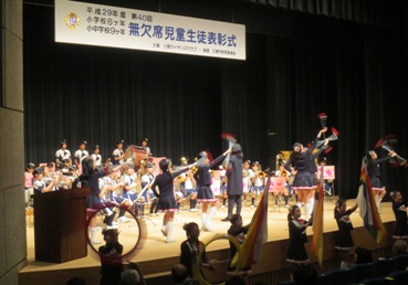 「平成29年度第40回小学校6ヶ年、小・中学校9ヶ年無欠席児童生徒表彰式」と書かれた横断幕が吊り下げられているステージ上で、バトントワリングを披露するユニフォームを着たバトンクラブの子どもたちと、その後ろに並べられたパイプ椅子に座り吹奏楽を披露する子どもたちの写真