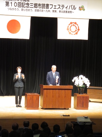 「第10回記念三郷市読書フェスティバル」の看板が掛かった舞台上で演台に立ち挨拶をする市長と、向かって左隣で手話通訳をする女性の写真