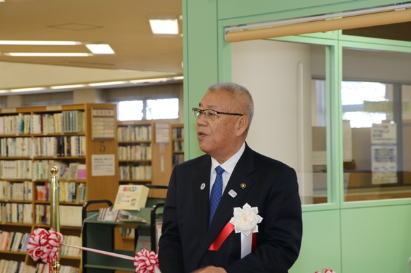 北部図書館に設置された「みる・よむ・きく バリアフリーコーナー」の薄緑の壁の前で、胸章を付けた三郷市長が挨拶の言葉を述べている写真
