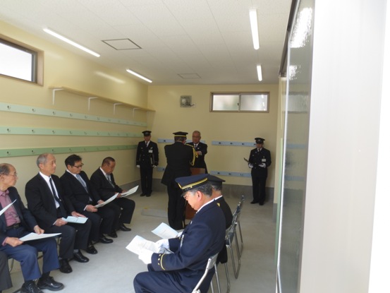 向かい合わせに設置されたパイプ椅子に関係者たちが座っている中、部屋の奥で市長から本間消防団長に目録を交付している様子の写真