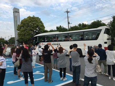 バスに乗り込んだ子供たちを駐車場で手を振って見送っている保護者の後ろ姿の写真