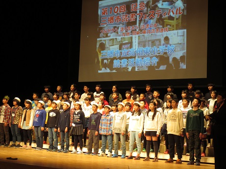 舞台上で、取り組み発表のスライドが映し出された後方のスクリーンの前に、たくさんの小学生が前後4列に並び正面を向いて立っている発表の様子写真