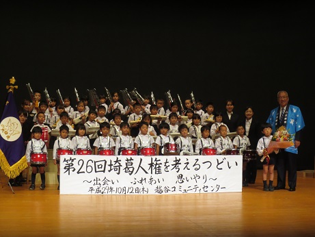 「第26回埼葛人権を考えるつどい」と書かれたボードの後ろにマーチングドラムや鍵盤ハーモニカを持った、制服姿の天使幼稚園の子どもたちが並んで立ち、最前列右端の園児が大きな花束を持ち、その隣に並ぶ青い法被を着た三郷市長とともに撮影した記念写真