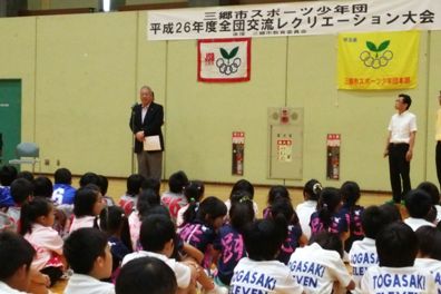 後ろの壁に「三郷市スポーツ少年団平成26年度全団交流レクリエーション大会」と書かれた横断幕の手前に設置されたマイクスタンドの前に立ち挨拶を行っている市長の話を体育座りをして聞いている子供たちの後ろ姿の写真