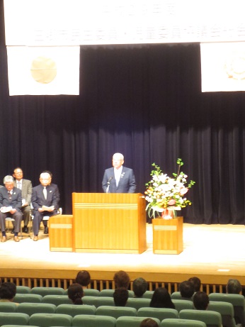 ステージ上に吊るされた「平成29年度三郷市民生委員・児童委員協議会総会」と書かれた横断幕に国旗と三郷市旗が貼り付けられており、鮮やかな花で彩られた花台と並ぶ演台に立ちあいさつの言葉を述べる三郷市長とステージ上に配置された席に座って話を聞く関係者の写真