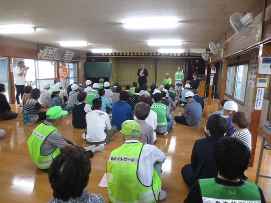 公民館内で、黄色のパトロールのベストを着用した参加者やヘルメットを被っている参加者たちが座って見守る中、正面に立って話をしている市長の写真
