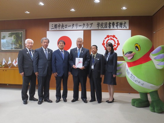 「三郷中央ロータリークラブ学校図書費寄贈式」と書かれた横断幕と国旗・三郷市旗が吊り下げられている壁の前で、祝儀袋を持った三郷市長が三郷中央ロータリークラブの関係者と白いタスキをかけたかいちゃんとともに記念撮影をしている写真