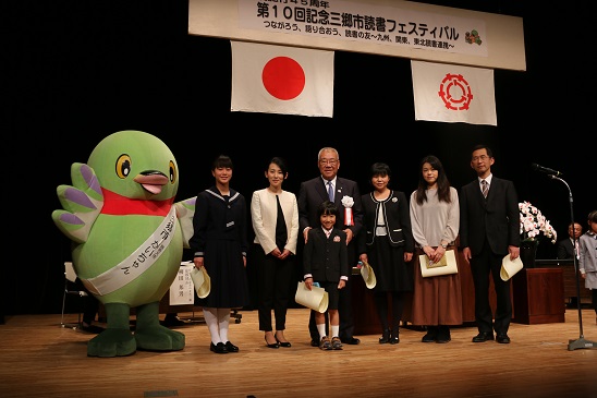 舞台上で、市長を中心に並んで立つ賞状を手にした6人の大人、子供と、向かって左に立つかいちゃんを撮影した記念写真